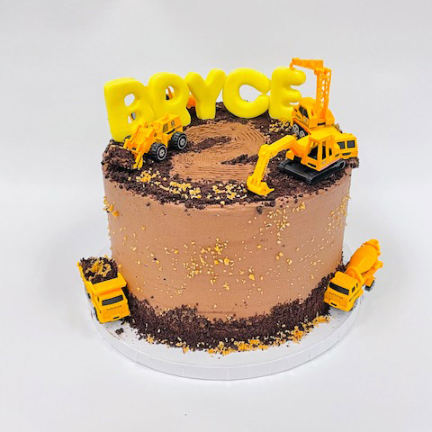 a singled tier brown cake with crushed brown cookie crumbs to look like dirt along the base of the cake and on the top. Bottom base has a dump truck with its box bed filled with cookie crumbs and a cement truck. The top of the cake has the name Bryce carved in fondant icing standing up and the crane placing the E in place. A front loader, excavator and the number 2 scraped into the cookie crumbs on the top of the cake
