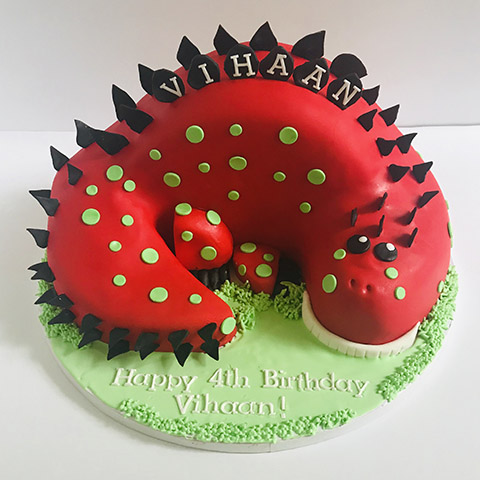 red dinosaur with green spots and black osteoderms is laying down on a green patch of grass in a c-shape design. Written on the grass in white icing, Happy 4th Birthday Vincent!