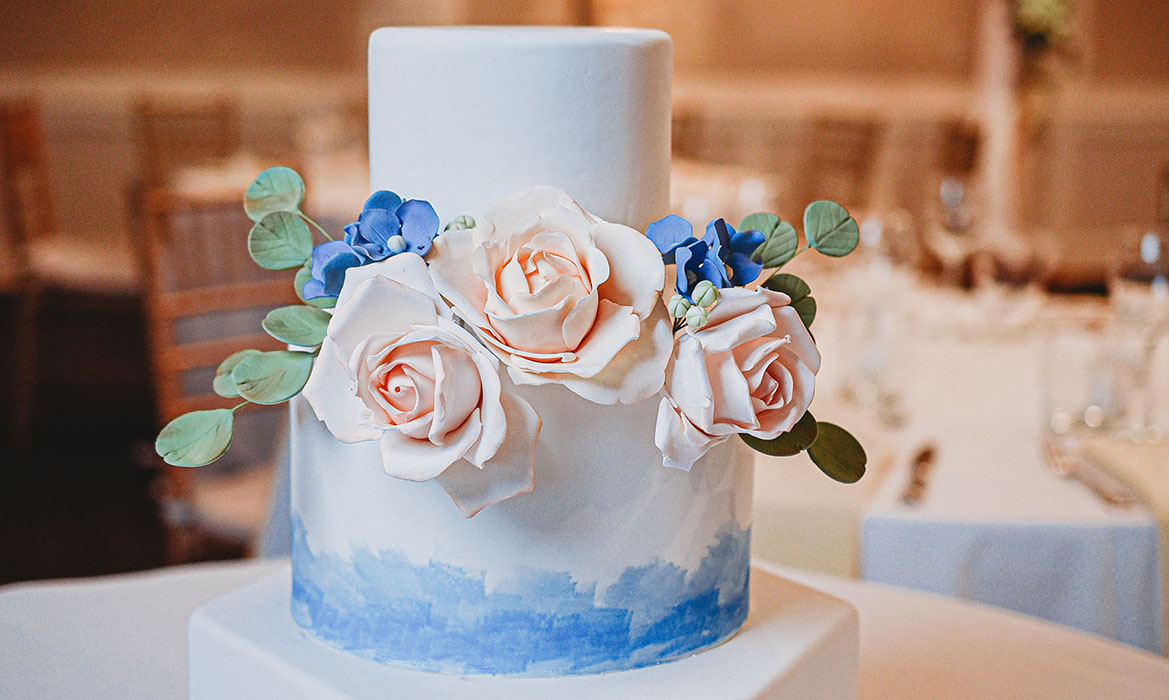 Deluxe wedding cake, two-teir with white smooth frosting and a hint of blue trim, three pink flowers made from icing, six little blue flowers made from icing, two sprigs of leaves made from icing